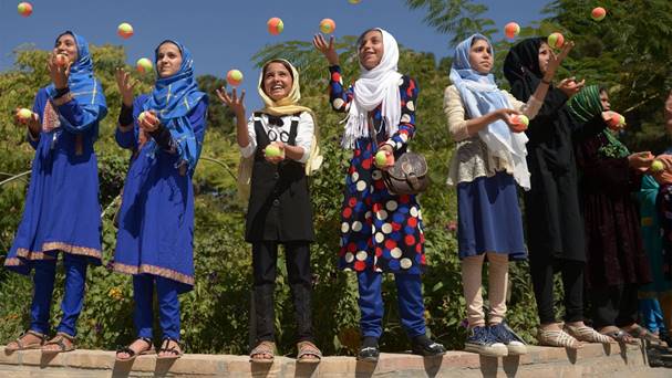 Several children stand on a low wall while they each juggle orange and yellow balls.