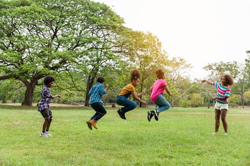 Two children hold the ends of a jump rope outside, while three others jump over the rope. 