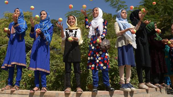 Several children stand on a low wall while they each juggle orange and yellow balls.