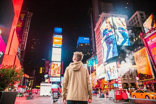 This is a picture of a man walking through Times Square in New York City. Around him, there are many different brightly lit advertisements for various products.