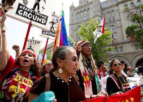 American Indian men and women, wearing traditional and modern clothing, hold signs that say, “keep it in the ground.” 